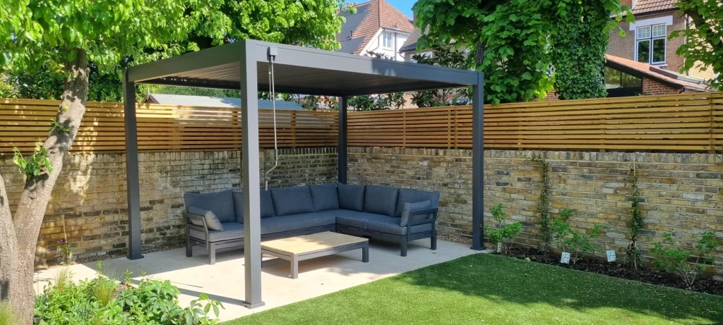 A modern garden seating area with a pergola, outdoor sofa and coffee table, set against a stone wall with planting and a lawn